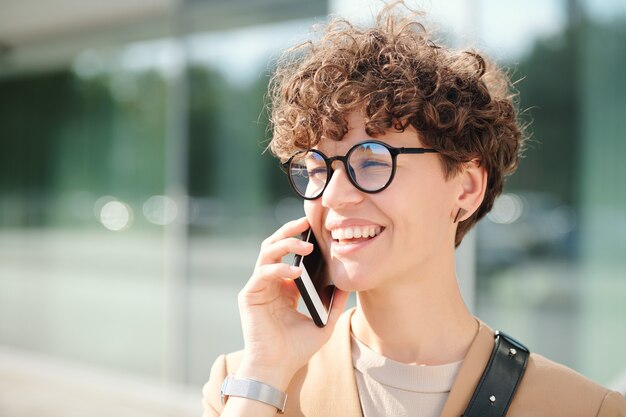 Felice giovane imprenditrice elegante con smartphone da orecchio consulenza client al telefono contro l'esterno del centro business contemporaneo