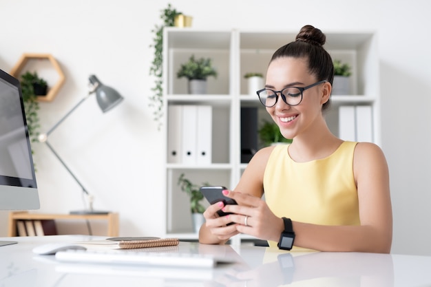 Felice giovane imprenditrice elegante con sorriso a trentadue denti scorrimento in smartphone mentre vi godete la pausa dal posto di lavoro