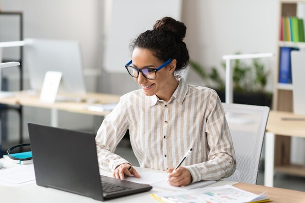 Felice giovane imprenditrice latina che lavora in ufficio seduto sul posto di lavoro utilizzando un computer moderno e prendendo appunti