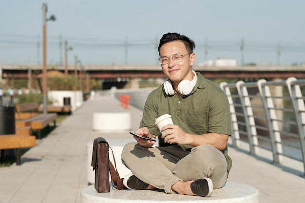 Felice giovane in abbigliamento casual che beve e scorre sullo smartphone in una calda giornata estiva mentre si riposa in un ambiente urbano