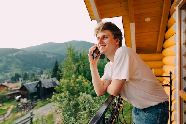 Felice giovane in abiti casual si trova sul balcone di una camera d'albergo sullo sfondo di un paesaggio di montagna e chiama al telefono distogliendo lo sguardo con un sorriso sul volto