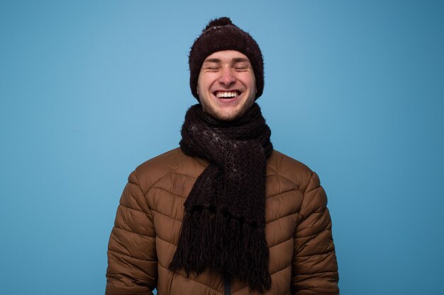 Felice giovane in guanti marroni e un cappello, giacca marrone chiaro ride su sfondo blu, ritratto in studio