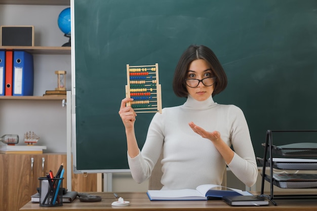 felice giovane insegnante femminile che indossa occhiali con in mano e indica l'abaco seduto alla scrivania con gli strumenti della scuola in classe