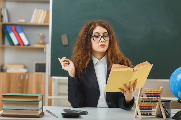 felice giovane insegnante femminile con gli occhiali che tiene e legge un libro seduto alla scrivania con gli strumenti della scuola in classe