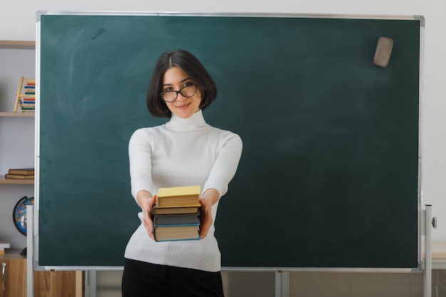 felice giovane insegnante femminile con gli occhiali che tiene libri fuori libri alla macchina fotografica in piedi davanti alla lavagna in classe