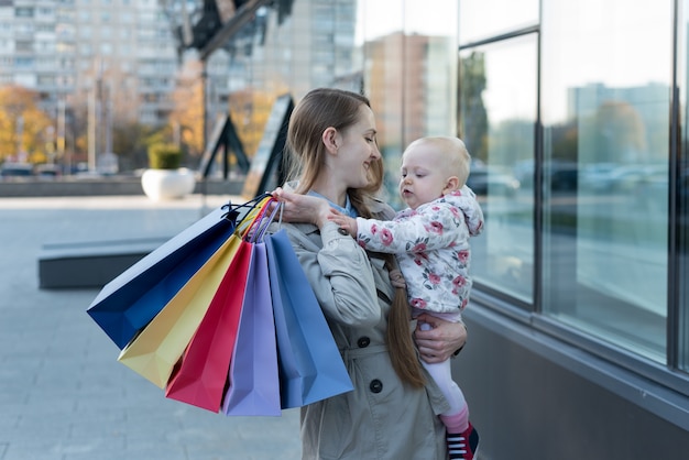Felice giovane madre con figlia piccola sulle braccia e borse della spesa in mano. Giornata di shopping.