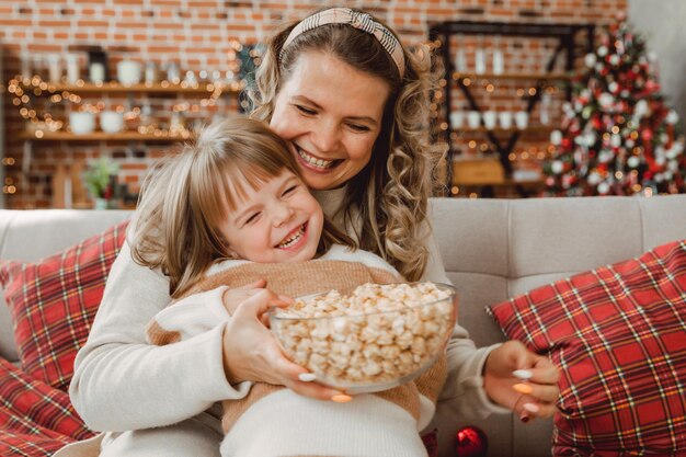 Felice giovane mamma e bambina ridono seduti sul divano e mangiano popcorn. famiglia che guarda la TV e mangia popcorn.