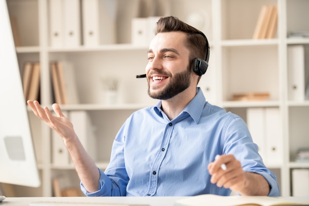 Felice giovane manager di ufficio con auricolare che spiega qualcosa a uno dei clienti durante la comunicazione tramite chat video