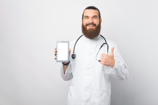 Felice giovane medico con la barba che mostra lo schermo vuoto del tablet e mostra il pollice in alto sul muro bianco