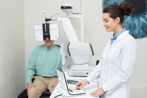 Felice giovane oculista femminile in camice bianco in piedi di fronte all'attrezzatura per il test della vista, guardando il display e girando la manopola