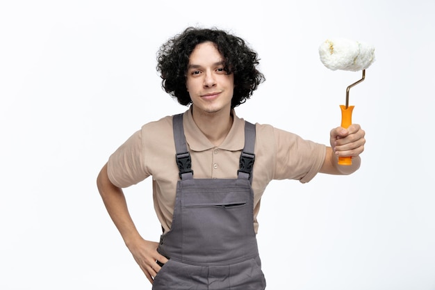 Felice giovane operaio edile maschio che indossa l'uniforme tenendo la mano sulla vita guardando la fotocamera che tiene il rullo di vernice isolato su sfondo bianco