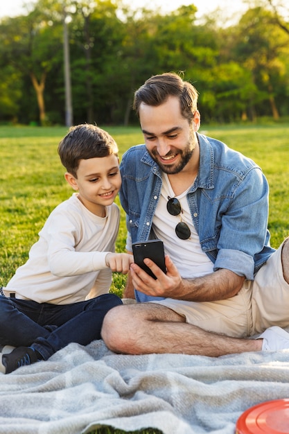 Felice giovane padre che fa un picnic con il figlioletto al parco, guardando il cellulare