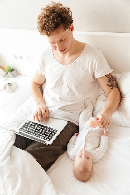 Felice giovane padre che gioca con il suo piccolo figlio mentre lavora su un computer portatile, sdraiato a letto