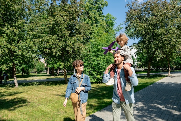 Felice giovane padre che trasporta carino piccolo figlio con aeroplano giocattolo sul collo e parlando con donna allegra mentre si cammina nel parco pubblico