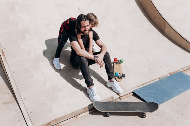 Felice giovane padre e suo figlio vestiti con eleganti abiti casual sono seduti in un abbraccio insieme sullo scivolo accanto agli skateboard in uno skate park nella calda giornata di sole.
