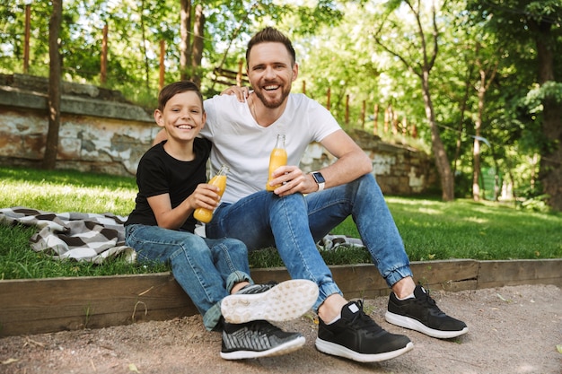 Felice giovane padre seduto con il suo piccolo figlio all'aperto a bere succo di frutta.