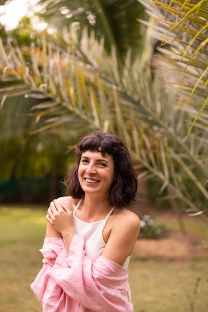 Felice giovane ragazza bruna caucasica in cima e camicia sorridente alla telecamera in piedi nel giardino tropicale Emozioni della gente stile di vita e concetto di moda