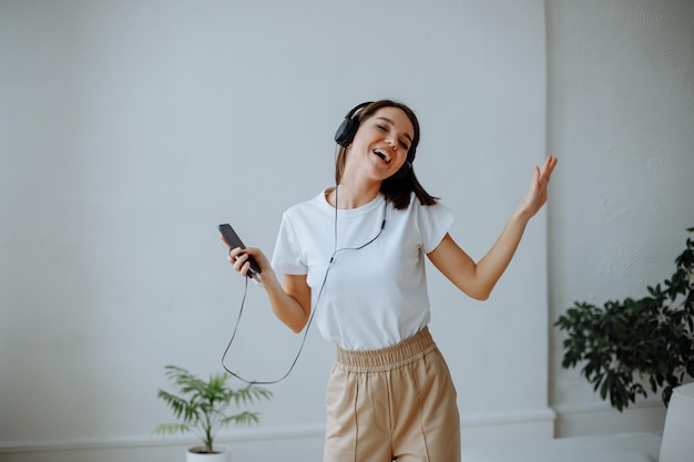 Felice giovane ragazza bruna in una maglietta bianca e jeans beige a casa ascoltando musica in cuffia