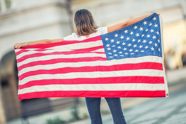 Felice giovane ragazza della scuola americana che tiene e sventola in città con la bandiera degli Stati Uniti.