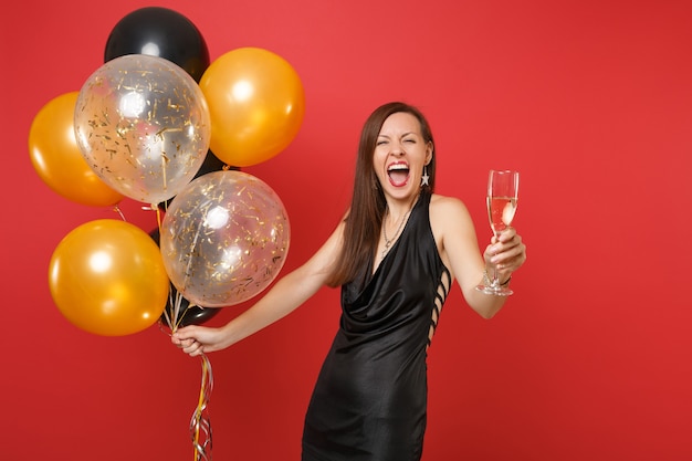 Felice giovane ragazza in abitino nero che celebra tenere un bicchiere di champagne palloncini d'aria isolati su sfondo rosso. Giornata internazionale della donna Felice Anno Nuovo compleanno mockup festa concetto.