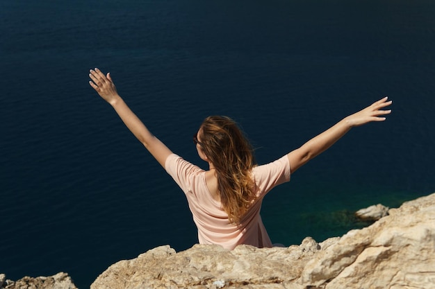 Felice giovane ragazza si siede su un'alta collina di pietra e gioendo alzando le mani sullo sfondo del mare e delle colline in una soleggiata giornata estiva