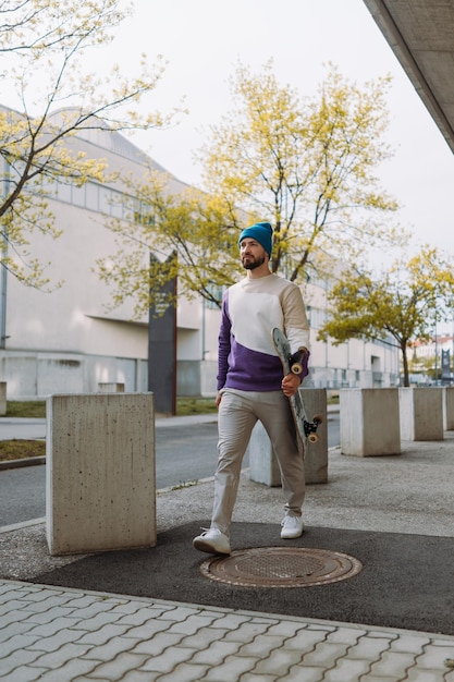 Felice giovane skater maschio in abbigliamento casual che trasporta skateboard mentre si cammina lungo la strada asfaltata foto verticale Foto di alta qualità