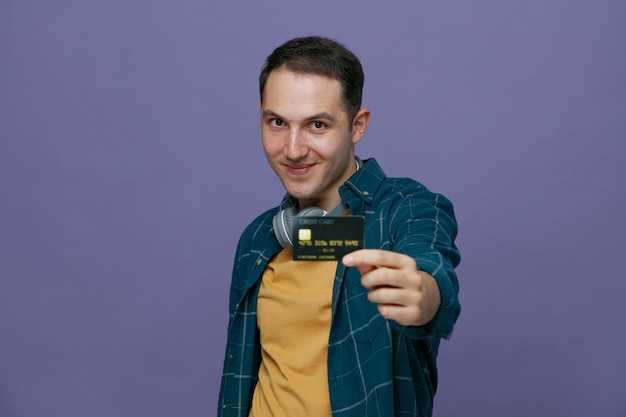 felice giovane studente maschio che indossa le cuffie intorno al collo in piedi nella vista di profilo guardando la fotocamera allungando la carta di credito verso la fotocamera isolata su sfondo viola