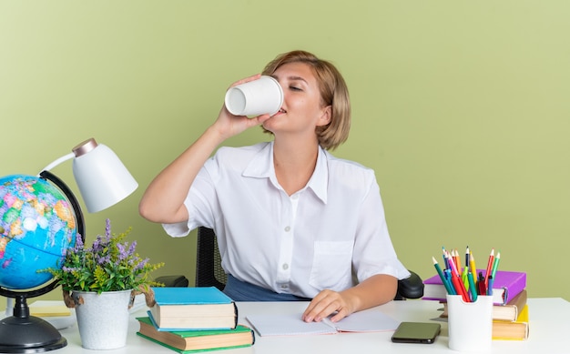 Felice giovane studentessa bionda seduta alla scrivania con gli strumenti della scuola che tiene la mano sulla scrivania bevendo caffè dalla tazza di caffè in plastica plastic