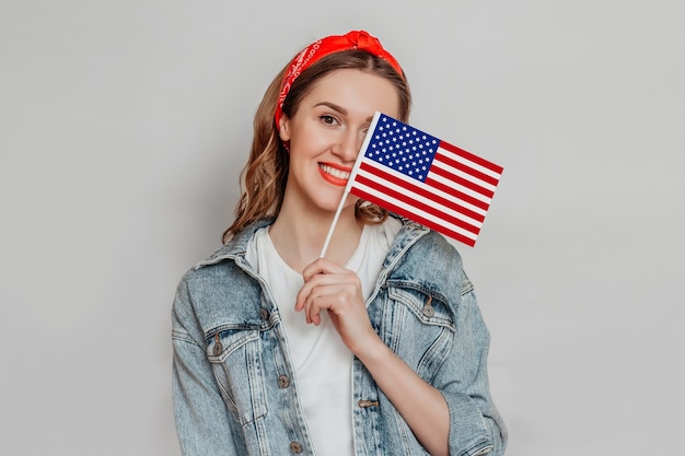 Felice giovane studentessa con rossetto rosso tiene una piccola bandiera americana e sorrisi isolati su sfondo grigio, ragazza con bandiera USA, 4 luglio giorno dell'indipendenza, spazio copia