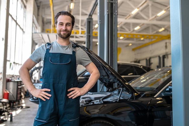 Felice giovane tecnico automobilistico caucasico vestito con abiti da lavoro in piedi in un'officina di riparazione auto