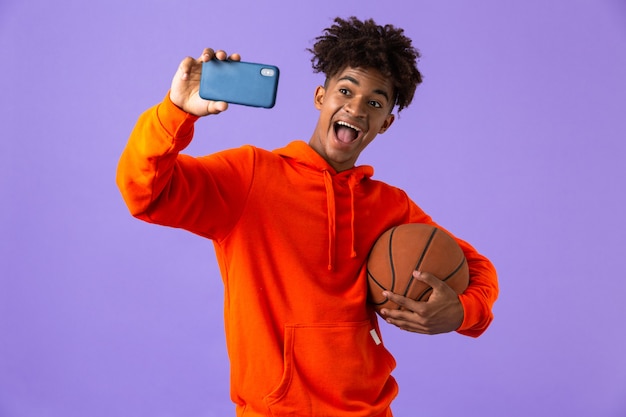 Felice giovane uomo africano in posa isolato su spazio viola prendere un selfie dal telefono tenendo la pallacanestro.