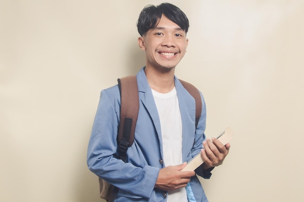 Felice giovane uomo asiatico che indossa un abito da college mentre trasporta borsa e libri su sfondo isolato