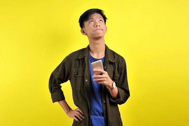 Felice giovane uomo asiatico che sorride usando lo smartphone guardando un'area vuota, isolata su sfondo giallo