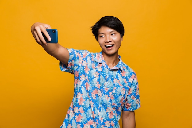 Felice giovane uomo asiatico in piedi isolato su uno spazio giallo prendere un selfie dal telefono cellulare.