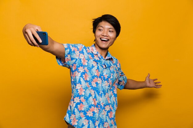 Felice giovane uomo asiatico in piedi isolato su uno spazio giallo prendere un selfie dal telefono cellulare.