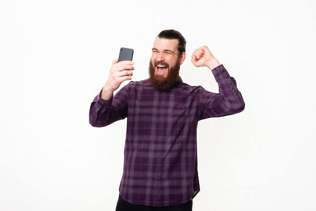 Felice giovane uomo con la barba guardando sorpreso smartphone e celebra la vittoria