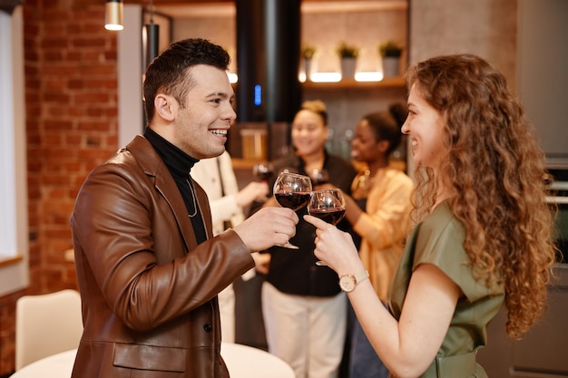 Felice giovane uomo e donna in abbigliamento casual intelligente che tintinnano con bicchieri da vino contenenti vino rosso e look