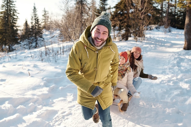 Felice giovane uomo in abbigliamento invernale che corre lungo la strada tra cumuli di neve mentre si tira la slitta con due ragazze gioiose e si diverte nella giornata invernale