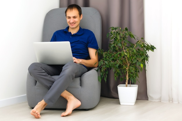 Felice giovane uomo in t-shirt seduto sul divano di casa, lavorando su un computer portatile, sorridente.