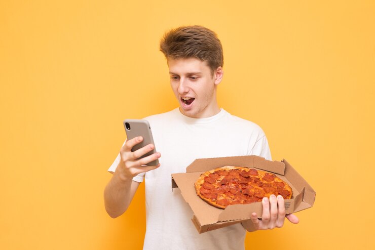 Felice giovane uomo in una maglietta bianca tiene una scatola di pizza e utilizza uno smartphone su un giallo