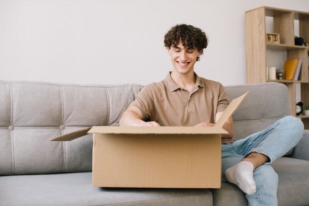 Felice giovane uomo riccio sorridente che apre la scatola con i regali di merci ordinate regali a casa sul divano Acquirente online cliente maschio che apre il pacco del negozio online