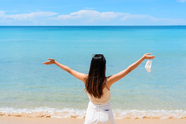 Felice giovane viaggiatore asiatico donna rilassante con maschera chirurgica sulla mano in spiaggia Nuovo stile di vita normale, assistenza sanitaria, viaggio e vacanza concetto Spazio libero