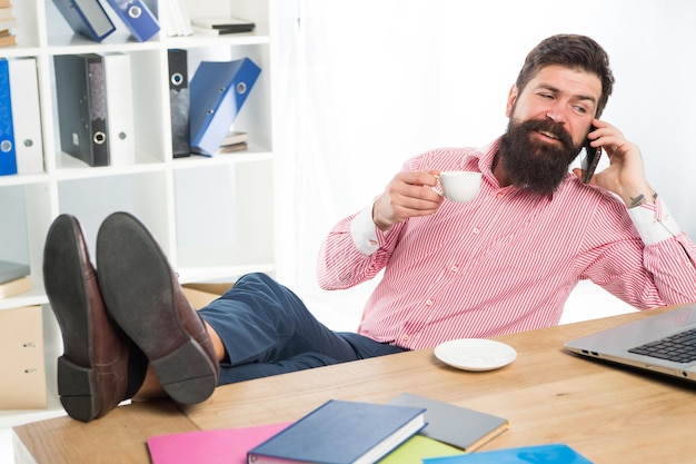 Felice hipster parla su smartphone bevendo tè in un ufficio moderno, conversazione.