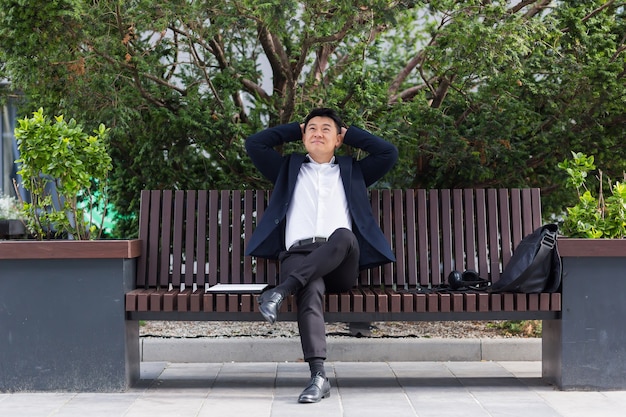 Felice impiegato asiatico, uomo d'affari seduto e riposato su una panchina nel parco cittadino all'aperto. Impiegato uomo d'affari un abito formale pausa riposo e relax dall'esterno. Strada del centro, fondo urbano