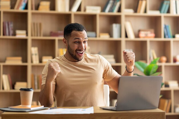 Felice imprenditore maschio arabo che utilizza il laptop gridando e gesticolando sì celebrando il successo aziendale seduto sul posto di lavoro