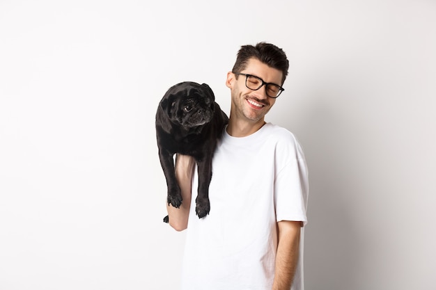 Felice l'uomo innamorato del cane, tenendo il cucciolo sulla spalla e sorridente, in piedi su sfondo bianco