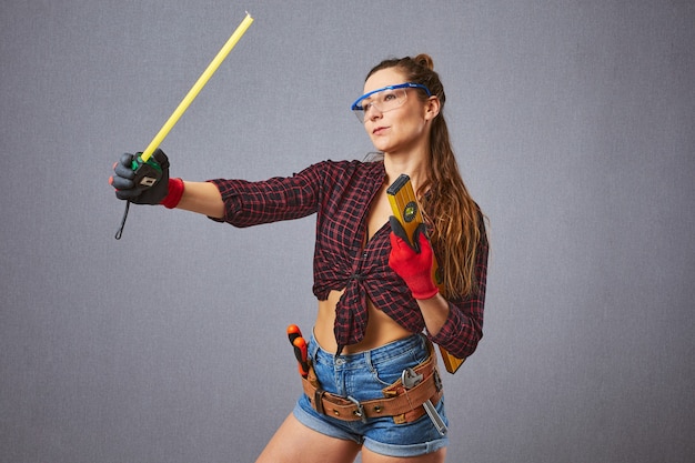 Felice lavoratore dai capelli lunghi che indossa occhiali protettivi. Misura di nastro della stretta della lavoratrice sexy. Riparazione, costruzione e concetto di costruzione - ragazza con strumenti di lavoro sul muro grigio.