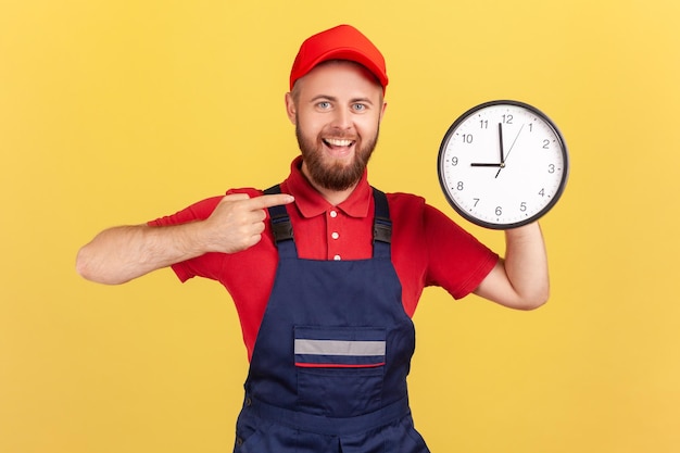 Felice lavoratore uomo in piedi e che punta al grande orologio da parete nelle sue mani guardando la fotocamera