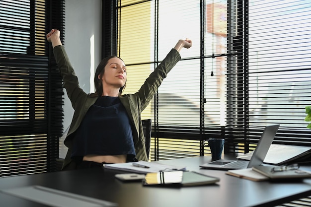 Felice lavoratrice caucasica che allunga le braccia rilassandosi sulla comoda sedia da ufficio nel moderno posto di lavoro dell'ufficio