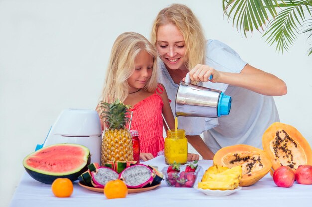 Felice madre bionda e sua figlia si divertono a preparare e bere frullati insieme a Bali. Famiglia caucasica in vacanza ropicale estiva bevendo succo di verdura verde fresco in un barattolo di frullato di vetro